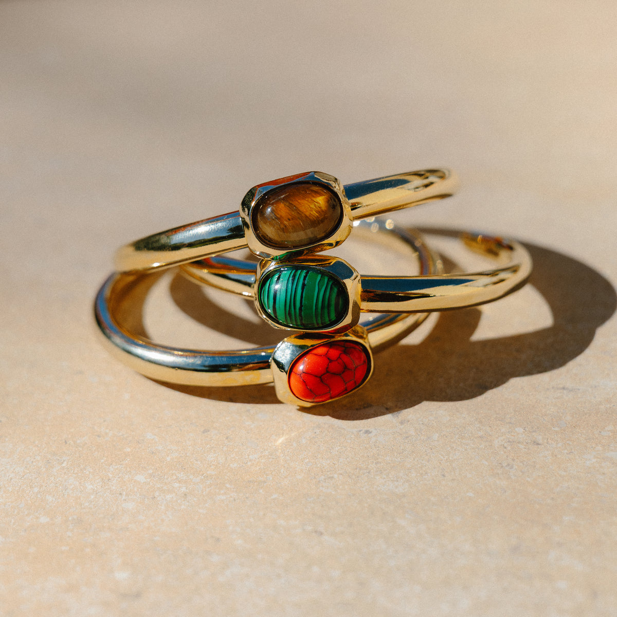 Tres brazaletes dorados Émolo con piedras ovaladas marrón, verde rayada y roja veteada, apilados sobre piedra al sol