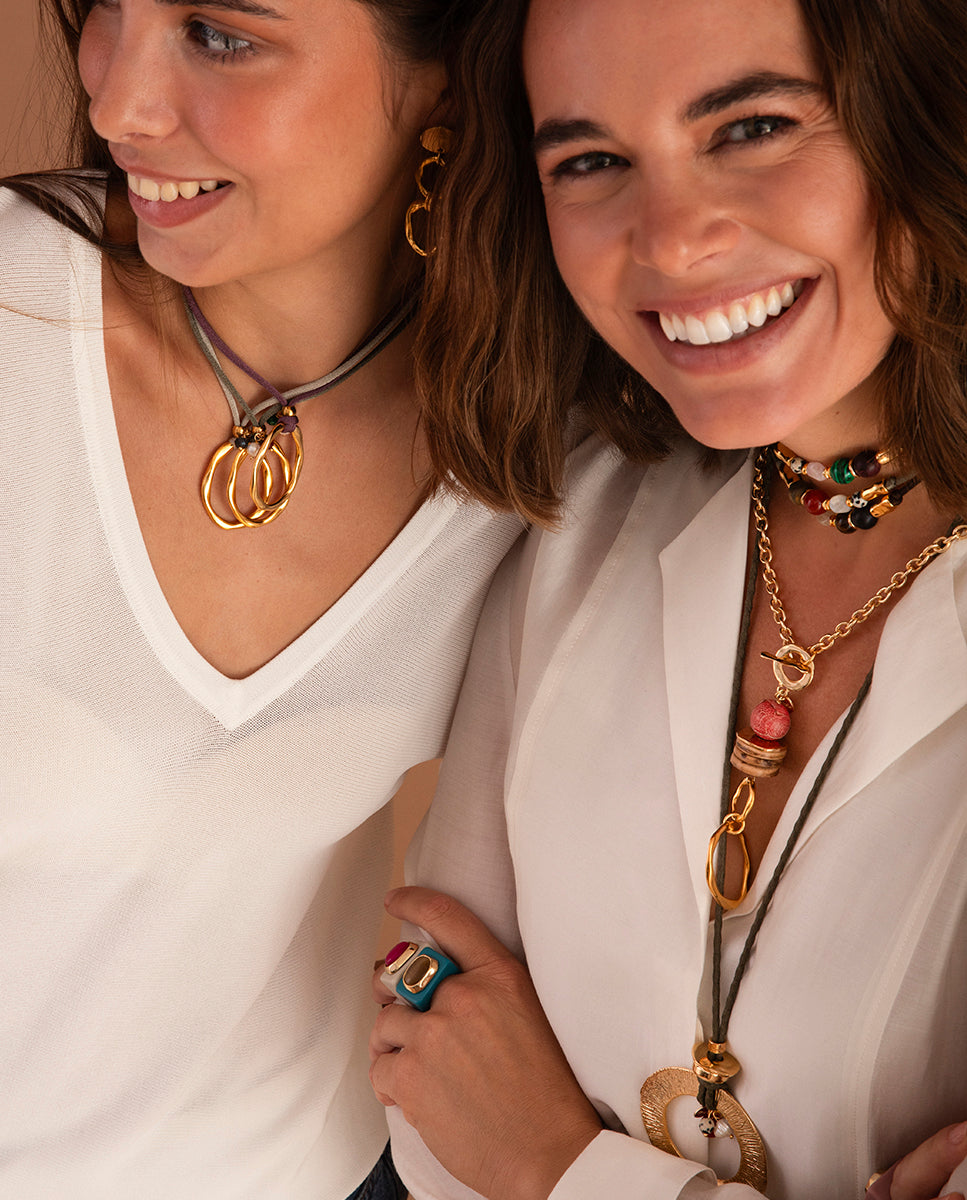 Primer plano de dos modelos con collares y pendientes dorados Emolo; en la mano se ven el anillo océano de selenita fumé y un anillo fucsia