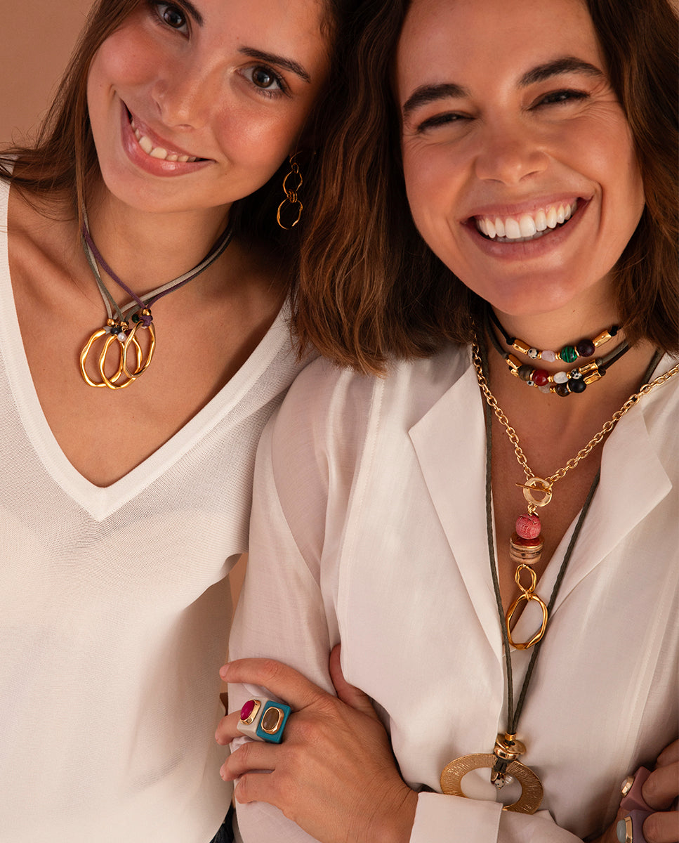 Dos modelos sonrientes con joyas Émolo; una luce el anillo océano de selenita fumé junto a un anillo fucsia, con collares de cordón y cadena dorada