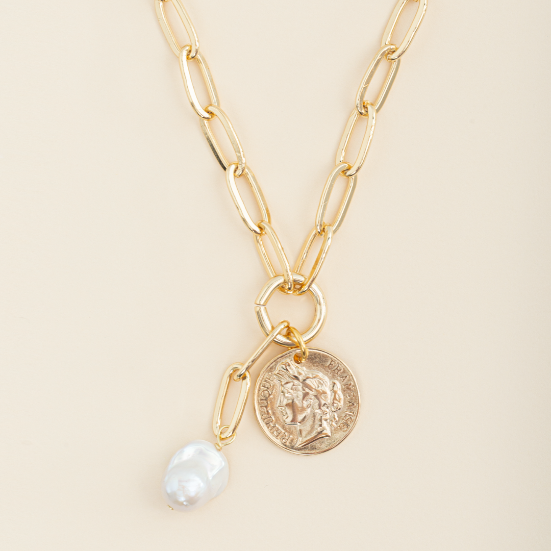 Detalle de collar de Émolo con cadena dorada de eslabones alargados, medalla redonda y perla blanca irregular